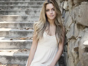 Photorealistic AI-generated blonde woman in a white summer dress sitting on stone steps in natural sunlight.