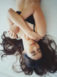 From an overhead view, a woman in a black bra lies on a bed with her eyes closed and arms crossed over her chest.