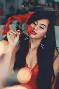 A woman with long dark hair holds a red rose to her face with her eyes closed.